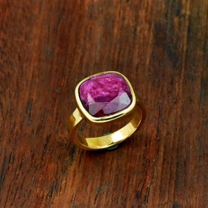 Op de afbeelding: Een gouden ring met een grote, vierkant geslepen, dieproze edelsteen met gefacetteerde oppervlakken. De ringband is glad en gepolijst en heeft een metallic gele afwerking die contrasteert met het levendige, getextureerde uiterlijk van de steen.