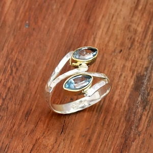 May include: A silver ring featuring two oval, light blue gemstones set in gold-coloured bezels. The ring has a hammered texture and a unique, flowing design, ideal for a jewellery collection.