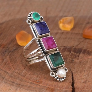 May include: A silver ring with four square gemstones. The stones are green, blue, pink, and white. The ring is set on a wooden surface.