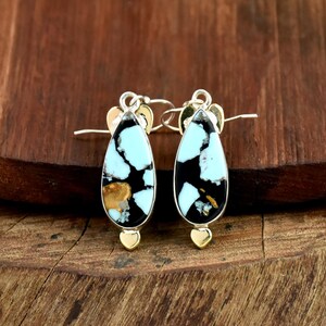 May include: A pair of teardrop-shaped earrings with a silver frame. The earrings feature a black background with turquoise and gold-colored stone inlays. The earrings have a gold-colored heart-shaped accent at the bottom and a silver hook.