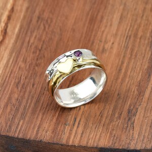 May include: A silver and gold-tone ring with a heart and a small purple gemstone. The ring has a spinning band and is set on a wooden surface. The heart is gold-colored, and the gemstone is round.