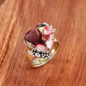 May include: A heart-shaped ring featuring a red, pink, and gold stone set in silver. The ring has a gold band and is adorned with small silver beads.