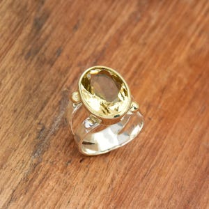 Puede incluir: Un anillo de plata con una gran piedra preciosa amarilla de talla ovalada engastada en un bisel dorado. El anillo tiene un diseño de caña dividida con pequeños acentos decorativos. El anillo se exhibe sobre una superficie de madera.