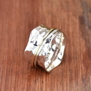 Puede incluir: Un anillo giratorio de plata y oro con textura martillada. El anillo está hecho de plata de ley y tiene una banda dorada alrededor del centro.