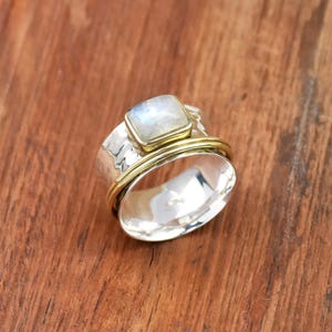 Op de afbeelding: Een zilveren ring met een vierkante, iriserende edelsteen gezet in een gouden bezel. De ring heeft een brede band met een gehamerde textuur en een gouden band rondom de edelsteen. De ring wordt getoond op een houten oppervlak.