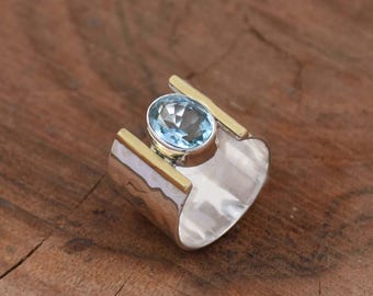 May include: A silver ring with a light blue gemstone set in a gold band. The ring has a hammered texture and a wide band.