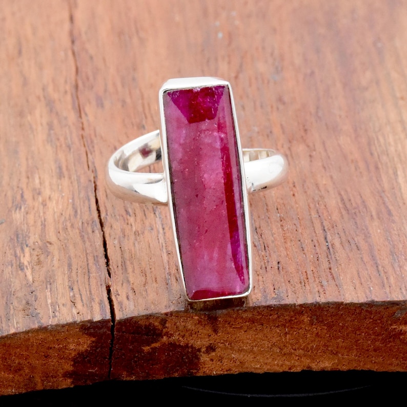 Op de afbeelding: Een zilveren ring met een rechthoekige, dieprode edelsteen. De edelsteen is gezet in een bezel setting en de ring heeft een eenvoudig, elegant ontwerp. De ring wordt gepresenteerd op een houten oppervlak.