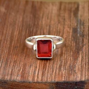 Pode incluir: Um anel de prata com uma pedra preciosa vermelha vibrante retangular. A banda do anel &eacute; lisa e polida, e a pedra preciosa &eacute; definida em uma moldura quadrada. O anel &eacute; exibido em uma superf&iacute;cie de madeira.