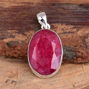 May include: A silver pendant featuring a large, oval-shaped ruby gemstone. The ruby is a deep red colour and has a faceted cut. The pendant is set in a silver bezel.