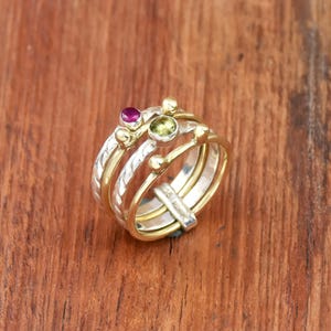 Könnte beinhalten: Ein Stapel von vier Ringen mit Gold- und Silberbändern. Zwei Ringe haben kleine, runde Goldakzente. Ein Ring hat einen kleinen, runden, magentafarbenen Edelstein, und ein anderer hat einen kleinen, runden, grünen Edelstein. Die Ringe liegen auf einer Holzoberfläche.