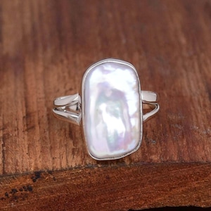 Könnte beinhalten: Rechteckiger Ring mit weißer Perle und Silberband. Die Perle hat eine schillernde Oberfläche. Der Ring liegt auf einer Holzoberfläche.