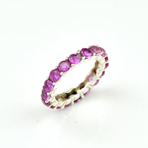 Peut inclure: Une bague en argent ornée d'une rangée continue de pierres précieuses roses vives. Les pierres sont de taille ronde et disposées côte à côte, créant un design d'éternité complet. La bague est présentée sur fond blanc.