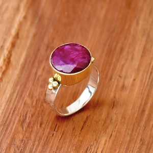 May include: A silver ring with a large, oval, deep pink gemstone set in a gold bezel. The band is silver with a textured finish and small gold accents near the gemstone. The ring is displayed on a wooden surface.
