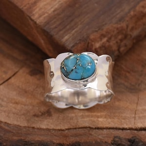 Può includere: Un anello d'argento con una grande pietra turchese ovale. La pietra ha una matrice di inclusioni color oro. L'anello ha una fascia larga con un bordo smerlato. L'anello è esposto su una superficie di legno.