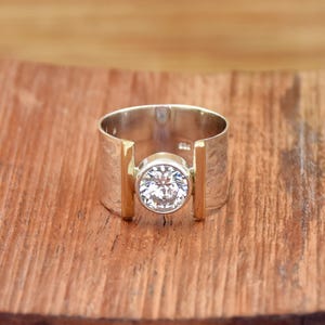 May include: A silver ring with a large, clear, round gemstone set in the center. The ring has a wide band with gold-colored accents on either side of the gemstone. The ring is displayed on a wooden surface.