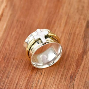 May include: A silver and gold spinning ring with a silver cross design. The ring is on a wooden surface.
