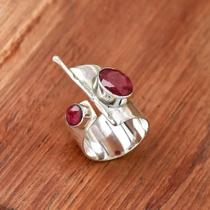 Puede incluir: Un anillo abierto de plata con dos piedras preciosas ovaladas de color rojo intenso engastadas en metal pulido. La banda tiene un diseño ancho y plano que termina en una punta en forma de hoja, con un acabado liso y brillante en toda la pieza.