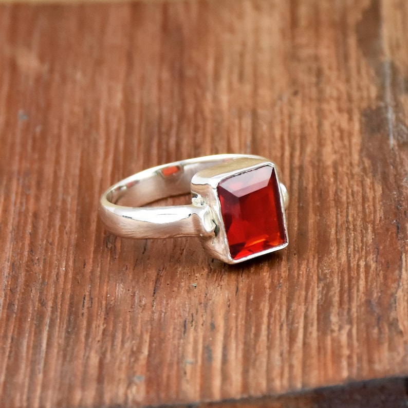 Pode incluir: Um anel de prata com uma grande pedra vermelha quadrada. O anel est&aacute; sobre uma superf&iacute;cie de madeira. A pedra preciosa &eacute; de uma cor vermelha profunda e vibrante, e a banda de prata tem um acabamento liso e polido. O anel &eacute; uma pe&ccedil;a marcante.
