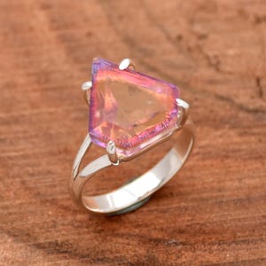 Puede incluir: Un anillo de plata con una gran piedra preciosa facetada e iridiscente. La piedra es de color rosa claro con un toque de morado.
