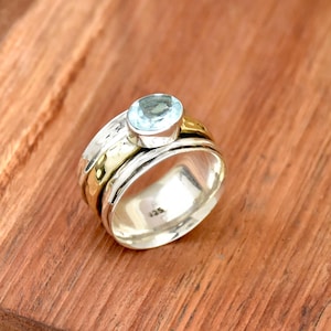 Op de afbeelding: Een draaibare zilveren en gouden ring met een lichtblauwe edelsteen. De ring ligt op een houten oppervlak.