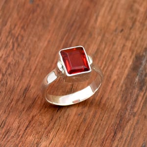 Può includere: Un anello d'argento con una pietra preziosa rettangolare di colore rosso intenso. La pietra preziosa è incastonata in una lunetta quadrata e l'anello ha una fascia semplice e lucida. L'anello è esposto su una superficie di legno.