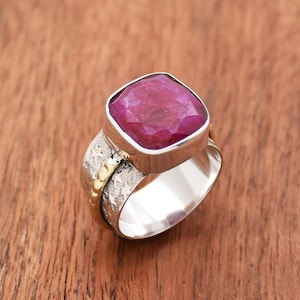 Puede incluir: Un anillo de plata con una gran piedra preciosa de rubí de forma cuadrada engastada en un engaste de bisel. La banda es texturizada y tiene una banda delgada de oro alrededor.