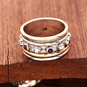 Puede incluir: Un anillo de plata y oro con una banda de tres filas. La fila superior tiene cinco piedras preciosas rojas y dos perlas blancas. El anillo está marcado con "125" en el interior.