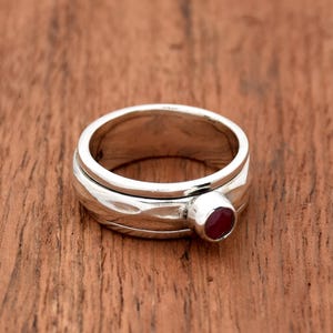 Könnte beinhalten: Ein drehbarer Silberring mit einem roten Edelstein. Der Ring besteht aus zwei Bändern, die sich unabhängig voneinander drehen.