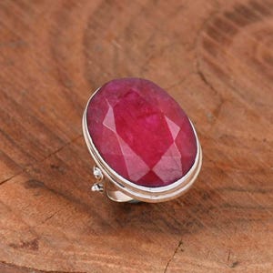 May include: A silver ring featuring a large, faceted, oval-shaped ruby gemstone. The ruby is a deep red colour.