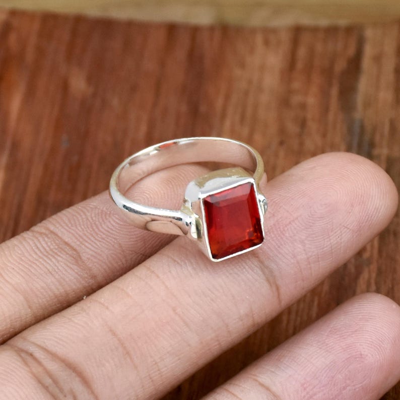Pode incluir: Um anel de prata com uma pedra preciosa retangular vermelha escura. O anel tem uma banda simples e uma moldura quadrada que segura a pedra vibrante. A cor da pedra preciosa contrasta com a prata.