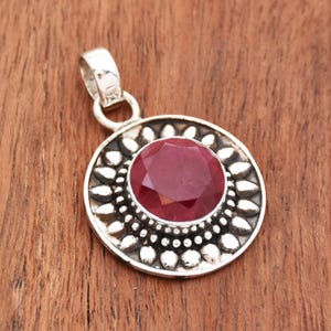 Peut inclure: Pendentif en argent avec une grosse pierre précieuse rubis rouge facettée au centre. Le rubis est entouré d'une monture argentée détaillée avec un motif floral. Le pendentif a une boucle pour une chaîne.