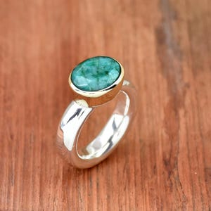 Puede incluir: Un anillo de plata con una piedra preciosa verde engastada en un bisel de oro. El anillo tiene una banda ancha y un acabado liso y pulido.
