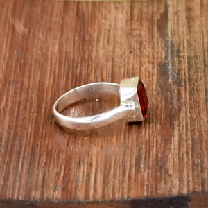 Pode incluir: Um anel de prata com uma grande pedra vermelha de corte quadrado. O anel est&aacute; sobre uma superf&iacute;cie de madeira, mostrando o metal polido e a cor vibrante da pedra. A banda do anel &eacute; lisa e simples.