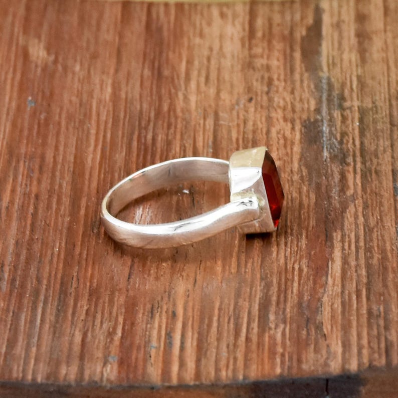 Pode incluir: Um anel de prata com uma grande pedra vermelha de corte quadrado. O anel est&aacute; sobre uma superf&iacute;cie de madeira, mostrando o metal polido e a cor vibrante da pedra. A banda do anel &eacute; lisa e simples.