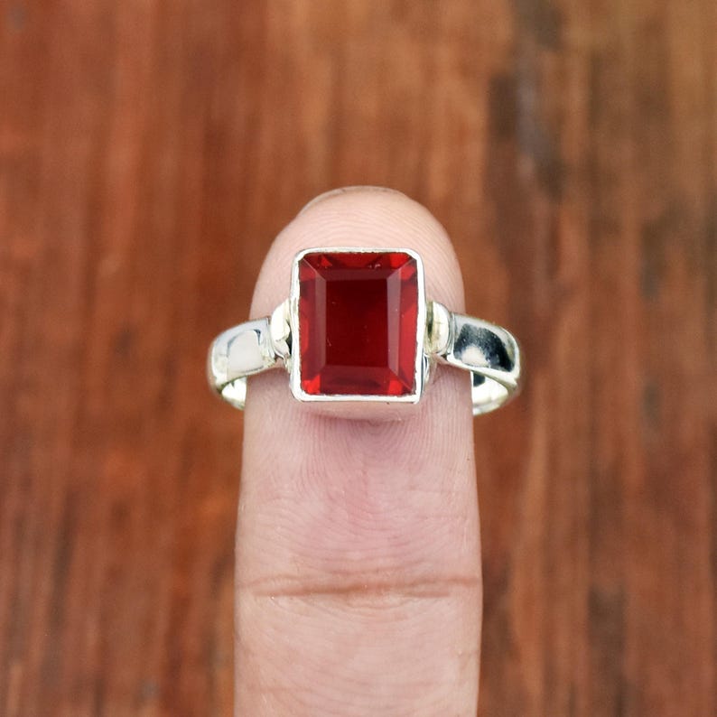 Pode incluir: Um anel de prata com uma pedra preciosa retangular, vermelho escuro. A banda do anel &eacute; prateada, e a pedra preciosa &eacute; colocada numa moldura quadrada. O anel &eacute; mostrado num dedo contra um fundo de madeira desfocado.