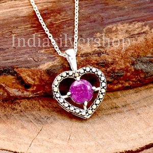 May include: A silver heart-shaped pendant necklace with a pink gemstone in the centre. The pendant is surrounded by small silver and gold beads.