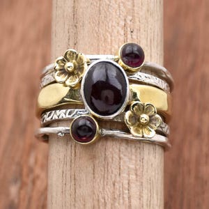 Könnte beinhalten: Ein Silber- und Goldring mit einem Stapel von Bändern, die mit drei kleinen roten Edelsteinen und einem größeren ovalen roten Edelstein verziert sind. Der Ring ist mit zwei kleinen Blumendesigns verziert.