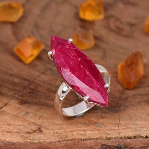 May include: A silver ring featuring a large, elongated, faceted ruby gemstone. The ruby is a deep, vibrant red color. The ring's band is a polished silver, and the gemstone is securely set with small prongs.