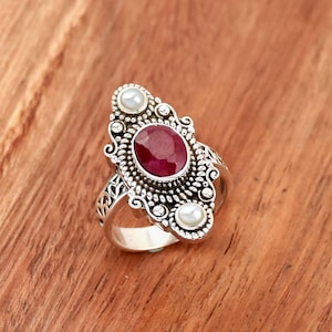 Peut inclure: Une bague en argent avec un design orné mettant en vedette une pierre précieuse de rubis rouge et deux perles blanches.