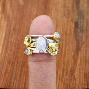 Puede incluir: Un conjunto de anillos de plata y oro con detalles florales y de piedras preciosas. Los anillos presentan una gran piedra lunar ovalada y detalles más pequeños de piedra lunar y flores. Se muestran en un dedo sobre un fondo de madera.