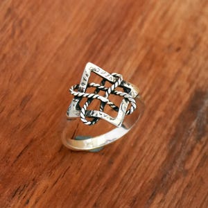 May include: A silver ring with a geometric design. The ring features a square-shaped frame with a woven rope-like pattern in the center. The ring is set against a wooden background.