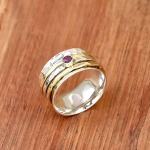 Può includere: Un anello color argento e oro con una piccola gemma rossa rotonda. L'anello ha una texture martellata sulla fascia argentata e fasce dorate lisce. L'anello è appoggiato su una superficie di legno.