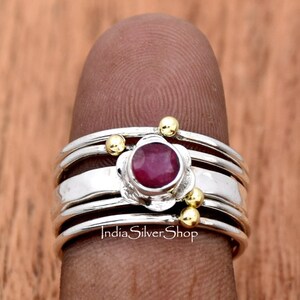 K&ouml;nnte beinhalten: Ein Silberring mit vier B&auml;ndern und einem roten Edelstein, der in einem Blumenmuster gefasst ist. Der Ring ist mit kleinen goldenen Perlen verziert.