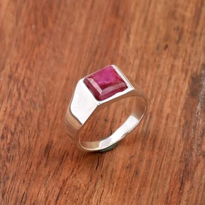 Op de afbeelding: Een zilveren ring met een vierkante, dieprode edelsteen. De ring heeft een gepolijste afwerking en een eenvoudig, modern design. De edelsteen is gezet in een vierkante bezel en de band is glad en afgerond.