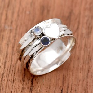 Op de afbeelding: Een zilveren draai ring met een hartvormige hanger en twee edelstenen. De ring heeft een gehamerde textuur en is bezet met een blauwe saffier en een maansteen.