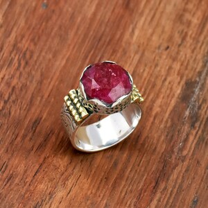 May include: A silver ring with a large, faceted, oval-shaped ruby gemstone. The ring has gold accents on the sides and a detailed silver band. The ruby is a deep red color, and the ring is set on a wooden surface.