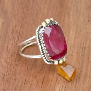以下が含まれることがあります： 銀のリングに、大きなファセットカットの赤い宝石が、銀と金のベゼルにセットされています。リングは繊細なねじれたバンドと、小さなぶら下がった琥珀色の宝石が付いています。