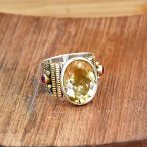 Könnte beinhalten: Ein silberner Ring mit einem großen, oval geschliffenen gelben Edelstein. Der Ring hat goldfarbene Akzente und kleine rote Edelsteine an den Seiten. Der Ring liegt auf einer Holzoberfläche.
