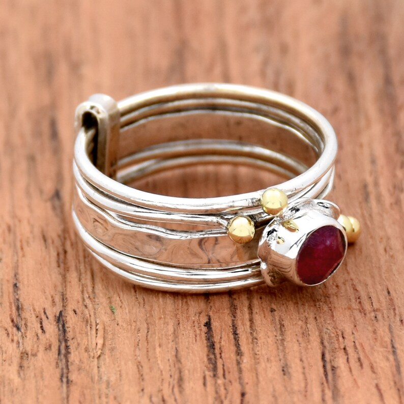 K&ouml;nnte beinhalten: Ein silberner Ring mit einem roten Edelstein und goldenen Akzenten. Der Ring besteht aus mehreren B&auml;ndern und hat eine geh&auml;mmerte Textur.