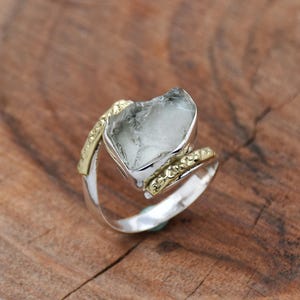 Könnte beinhalten: Ein silberner Ring mit einem großen, klaren, facettierten Edelstein, der in einer goldenen Fassung sitzt. Der Ring hat ein einzigartiges, gedrehtes Design.